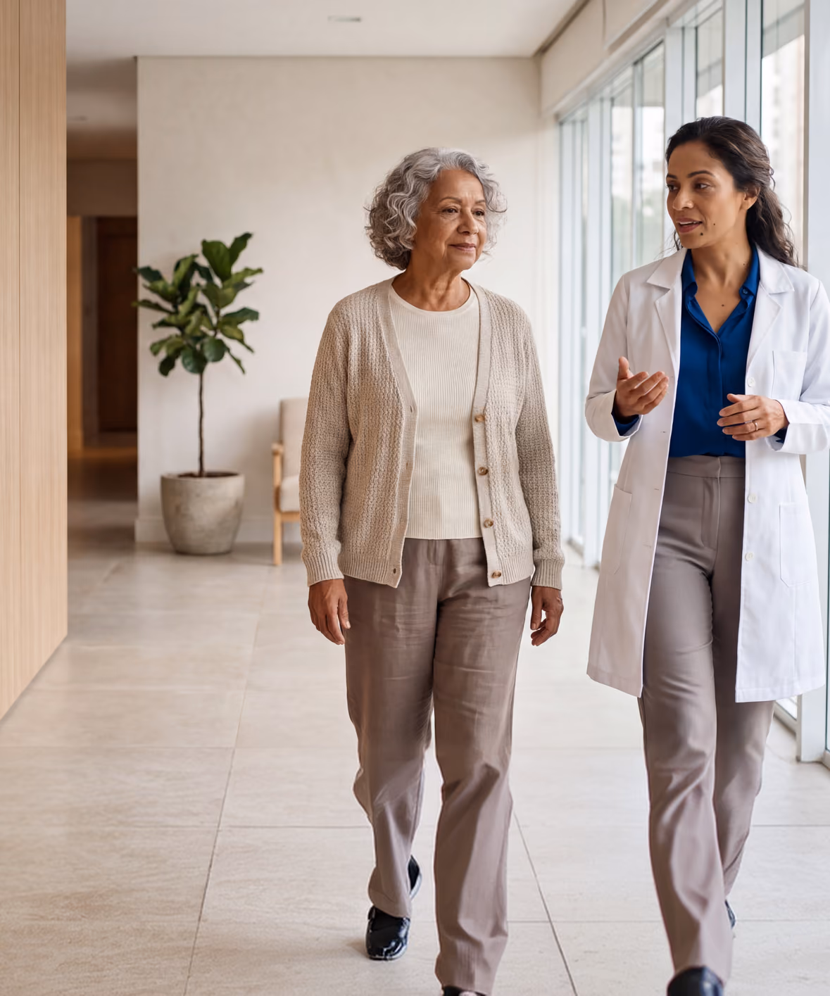 Médica caminhando e conversando com uma paciente idosa em um corredor iluminado
